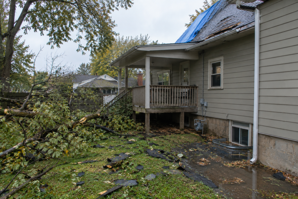 Storm-damaged Grand Rapids home with fallen limbs, scattered shingles, and a tarp covering part of the roof